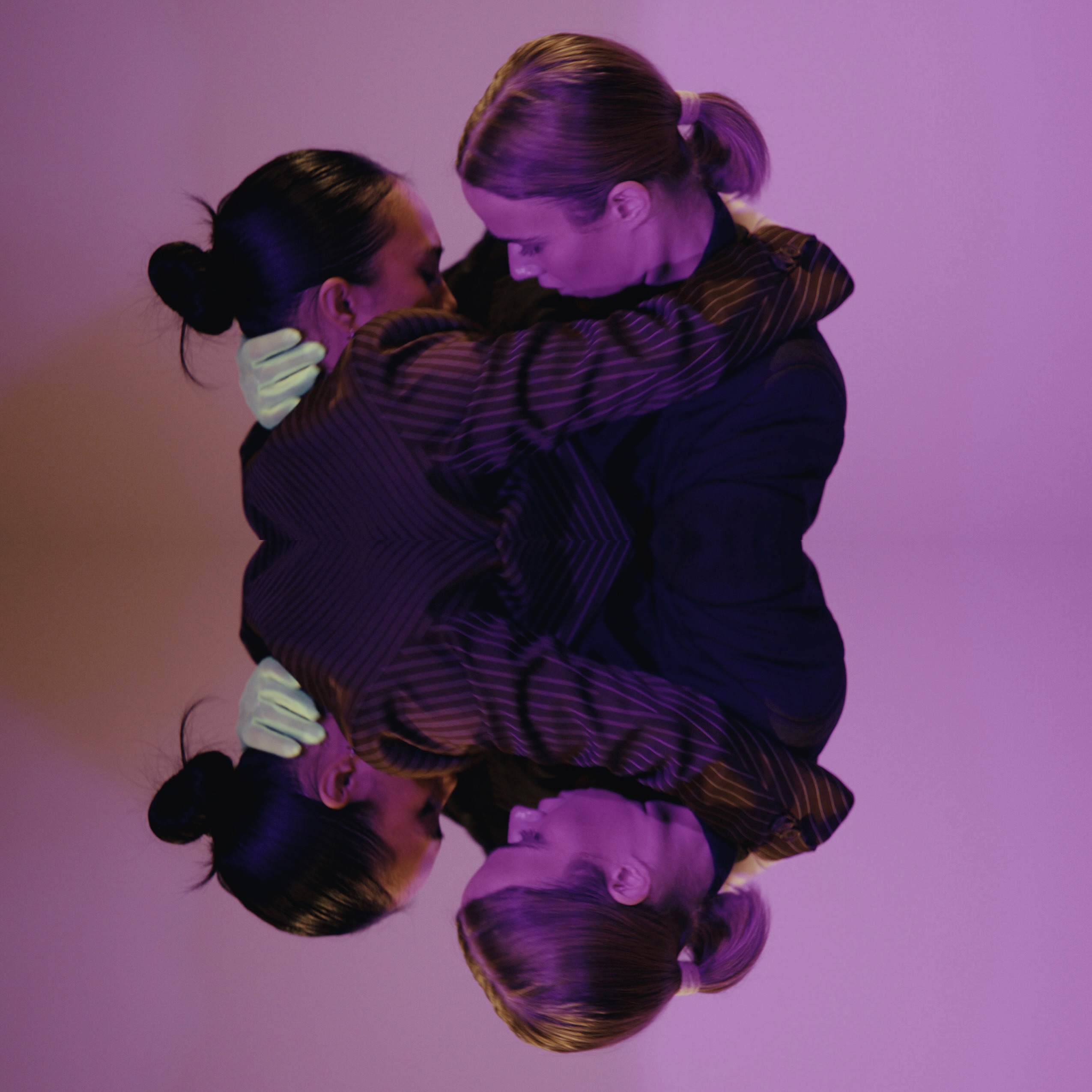 Two dancers, Kira Fargas and Hadassah Perry are wearing brown and grey suits and neon green gloves and are embraced in a hug with their faces close together. They are in front of a white wall, and are lit in purple lighting. The entire image is mirrored beneath them.