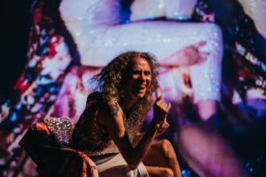 A light-skinned black woman in sequins is seated in front of a sequiny film still. Her hair is down, she is leaning forward, joyous, laughing. 