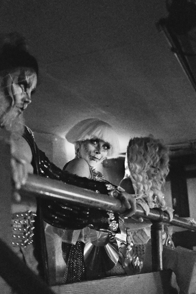 In this black-and-white image, three performers with dramatic makeup and costumes, including Lisa Frankenstein with a short white wig, lean against a railing, casting intense gazes that create a haunting, surreal atmosphere.