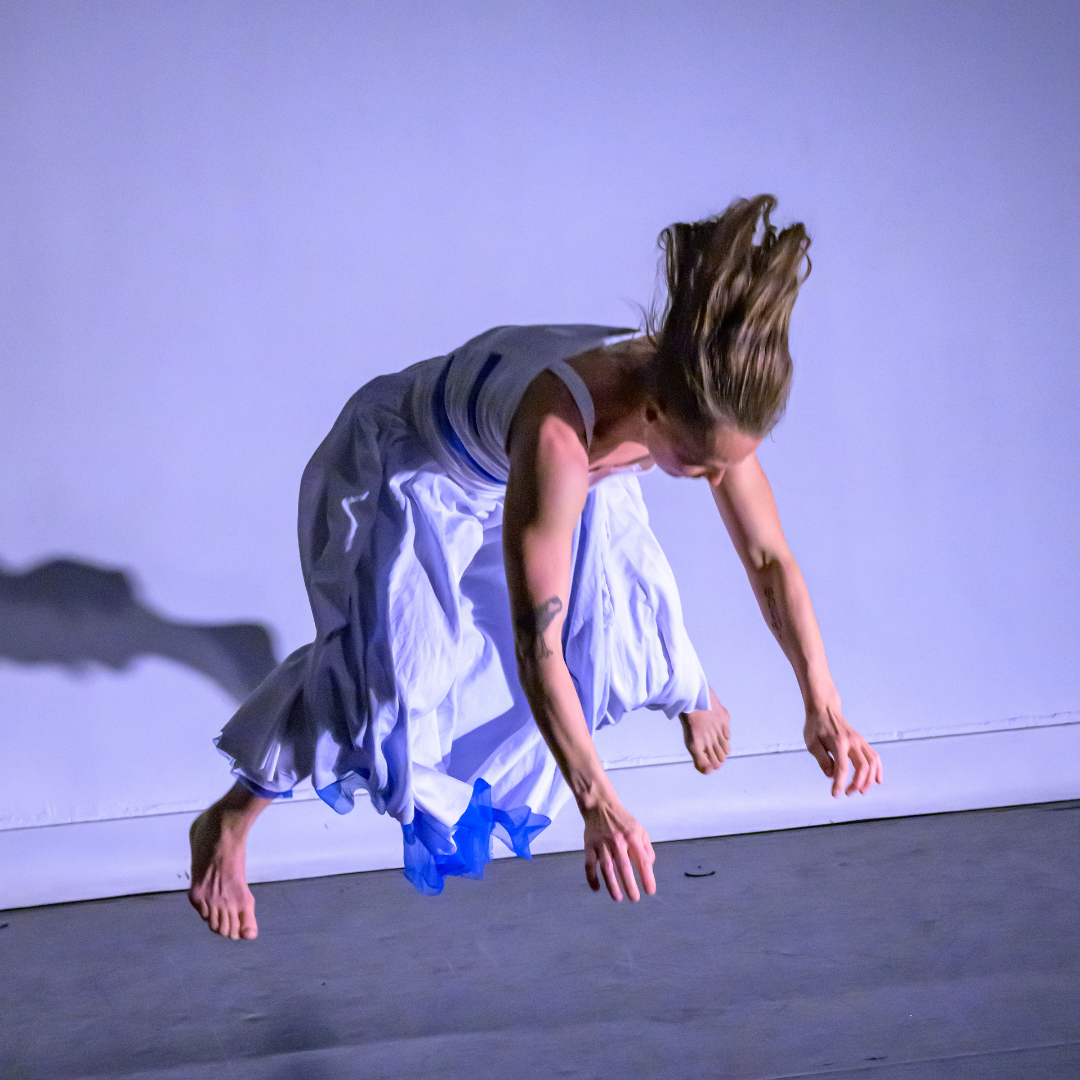 non binary dancer jumping in white top and skirt. Their focus in down and their body is parallel to the floor with arms and legs pointing downward 