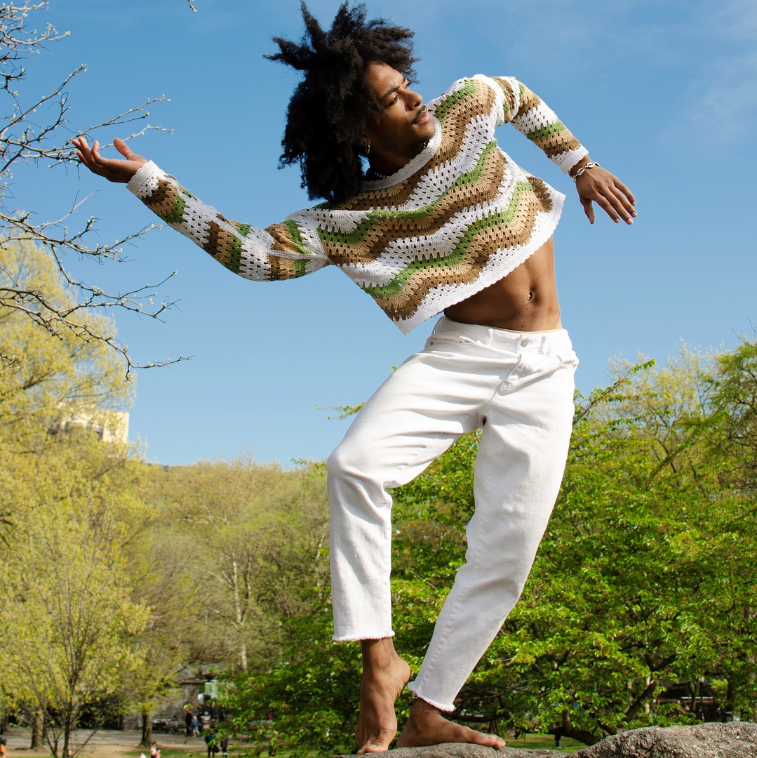 A male dancer in white pants and a striped sweater poses in front of a background of blue sky and green trees.