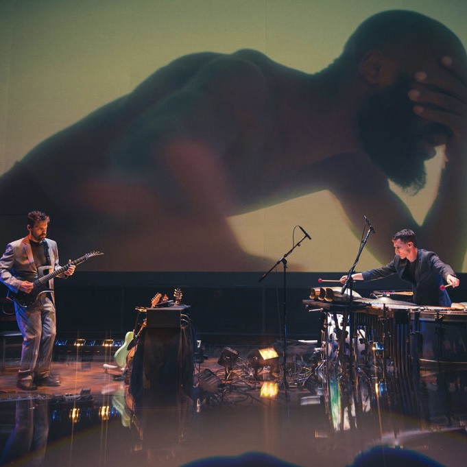 Two musicians stand apart with a movie screen behind them depicting a larger than life dancer leaning forward and covering his eyes. The musicians play an electric guitar, and percussion instrument.