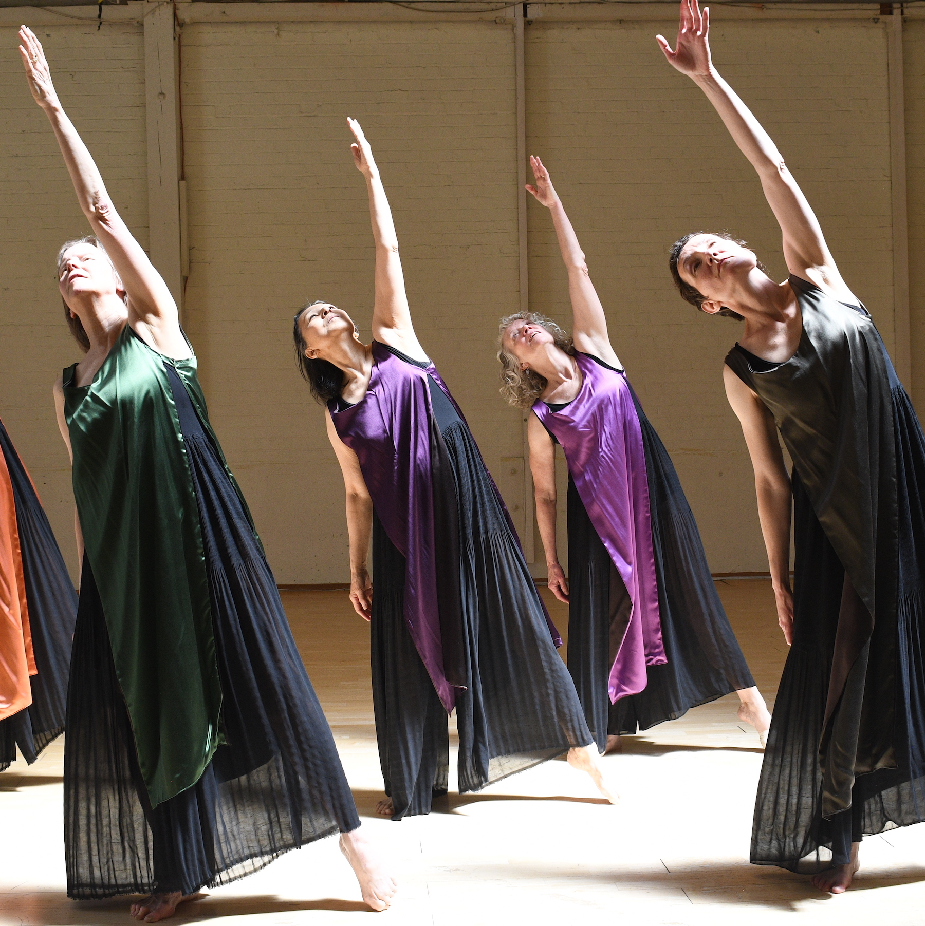 Four dancers dressed in long black tunics with accents of bright colored silks in purple and green are in a studio performance space with a hardwood floor. They reach their left arms long towards the sky with their gazes following. Their left legs are lengthened with toes touching the floor and their weight is shifted to the right.