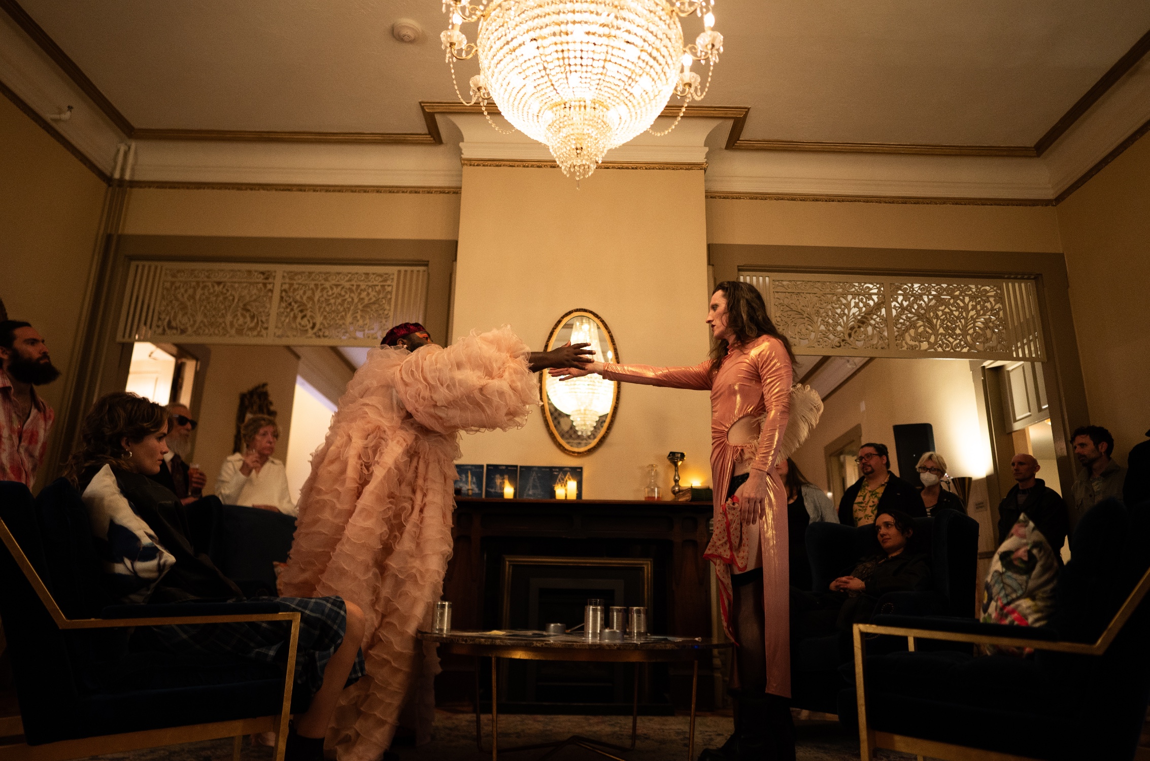 Two people wearing pink stand facing each other with an arm extended to the other. They are standing in a ballroom with a chandelier directly over their heads.