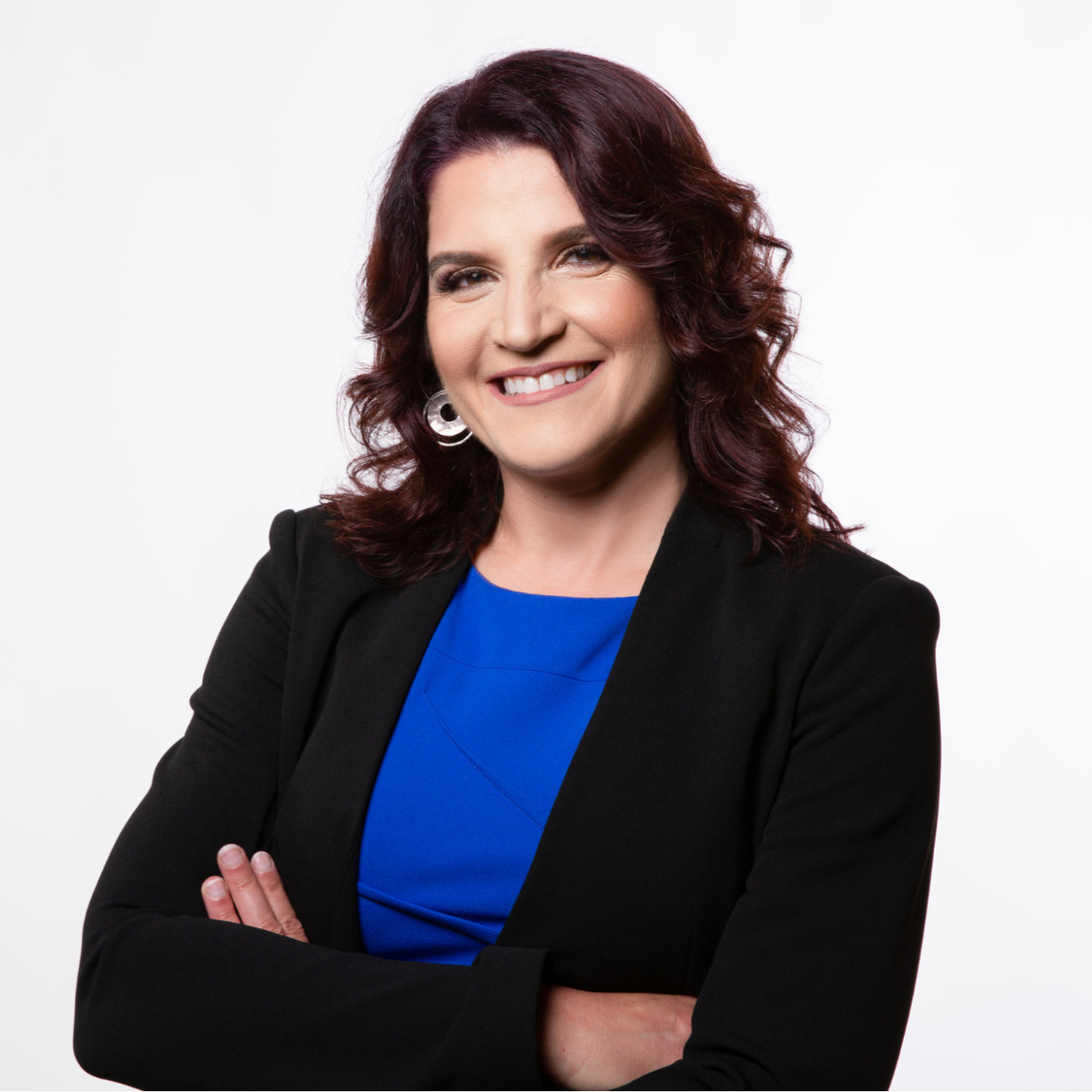 Image is a headshot of Stephanie Greenspan. The background is white. Stephanie has her arms crossed in front of her. She is wearing a black blazer and and bright blue top. She is wearing silver circle earrings and her hair is curly. Stephanie is smiling. 