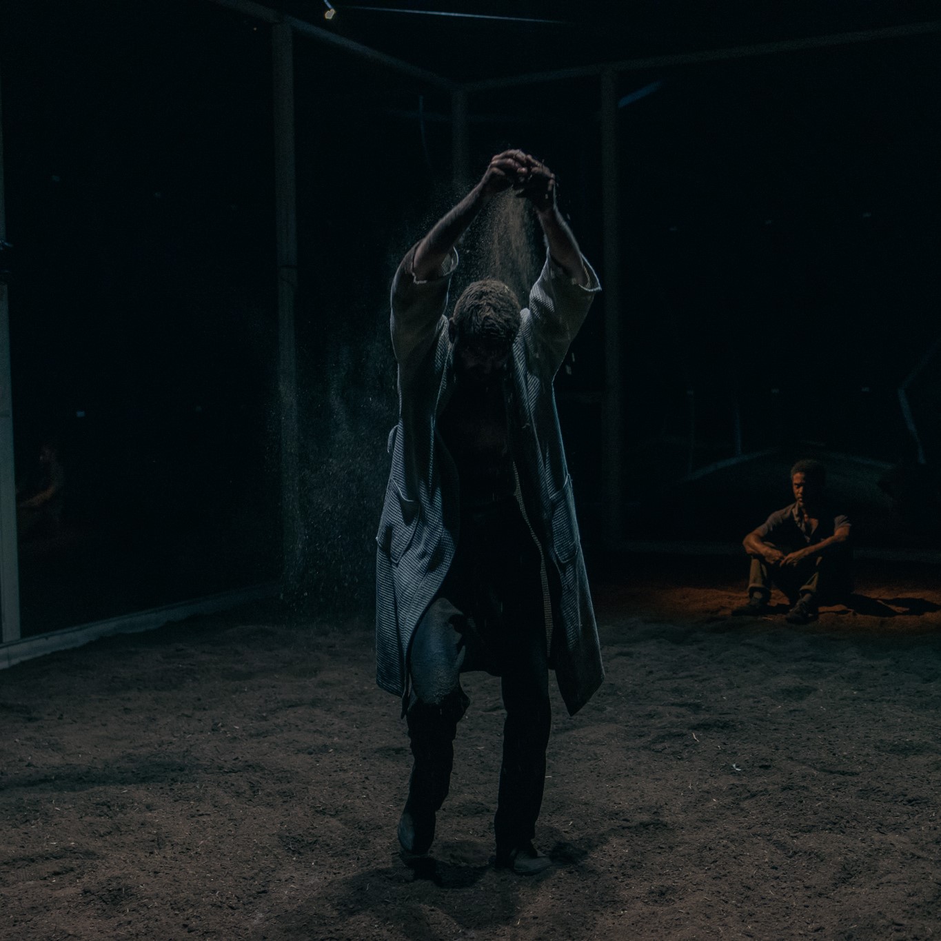 Two people are in a room, one is in the forefront of the image and the other is sitting in the back right. The one in the forefront is wearing a long coat and has their arms stretched above their head, dropping sand onto themselves. The entire room is dark and filled with sand.