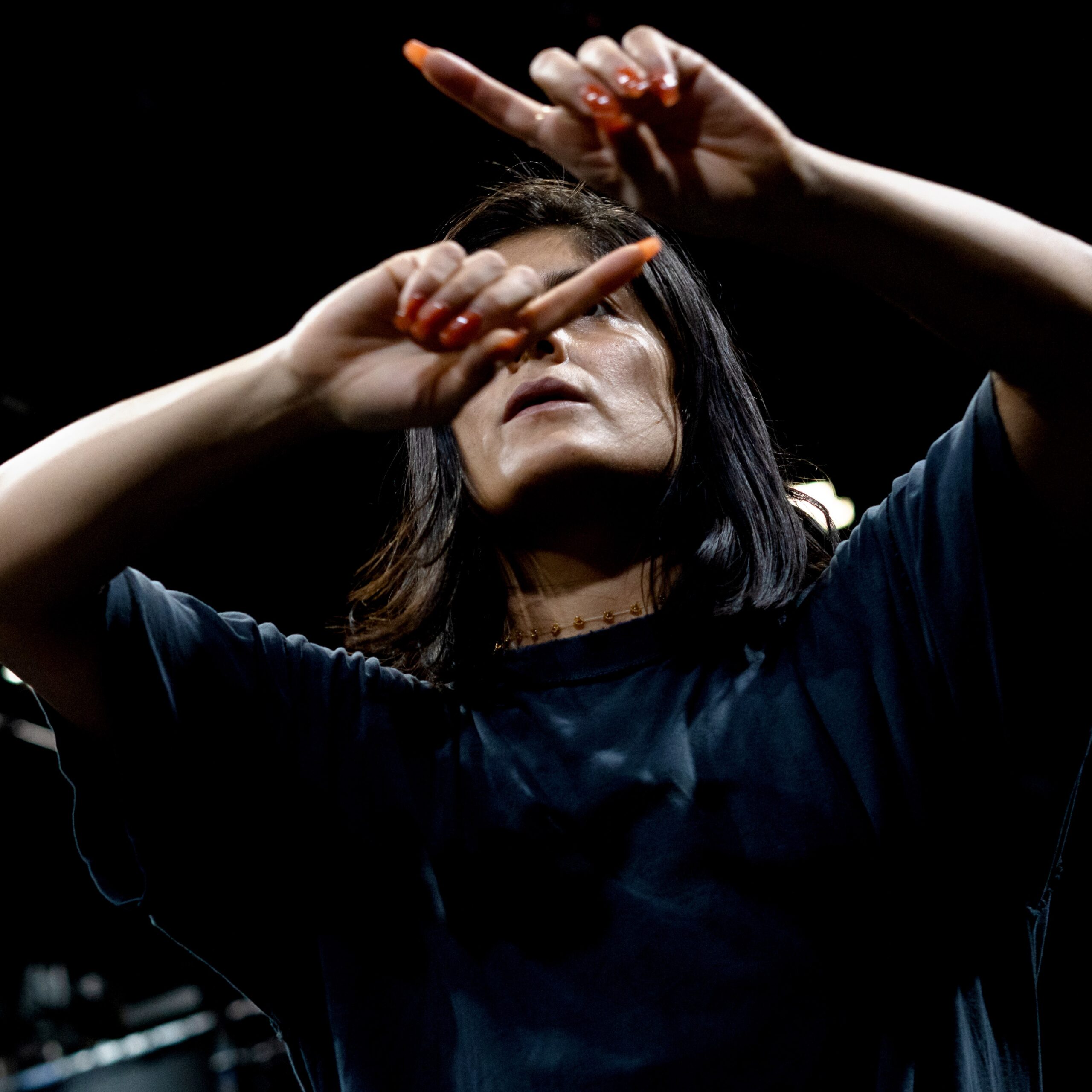 A person dances with two arms bent in front of their face, looking intentionally at their fingers with both pointer fingers outstretched. Their nails are long and a muted orange. They are wearing a baggy black t-shirt and an orange beaded necklace. Their hair goes to their shoulders and is straight and black. The background is black.