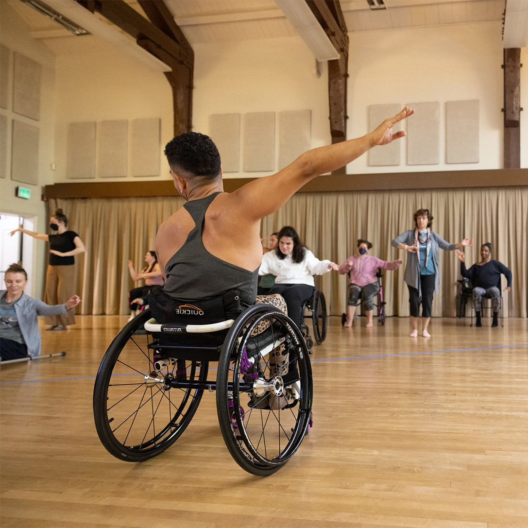 AXIS Dancer JanpiStar, who is a wheelchair user, teaches a class to a group of disabled and non-disabled dancers.