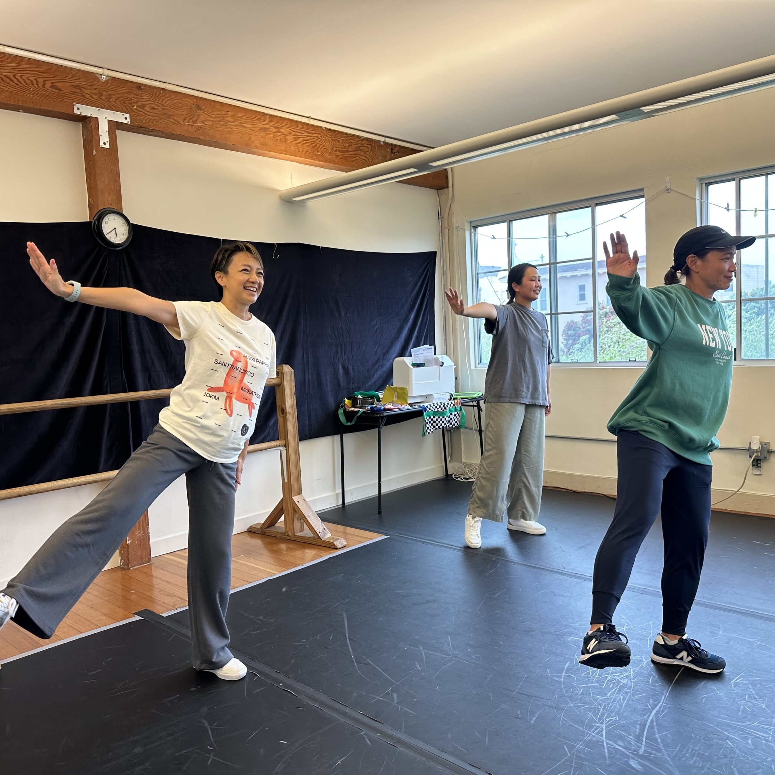 Three adult hip hop dancers wearing casual workout clothing and sneakers, all have one leg extended and one arm outstretched with a flexed hand. They are smiling as they watch their technique in mirror (not pictured) in front of them.