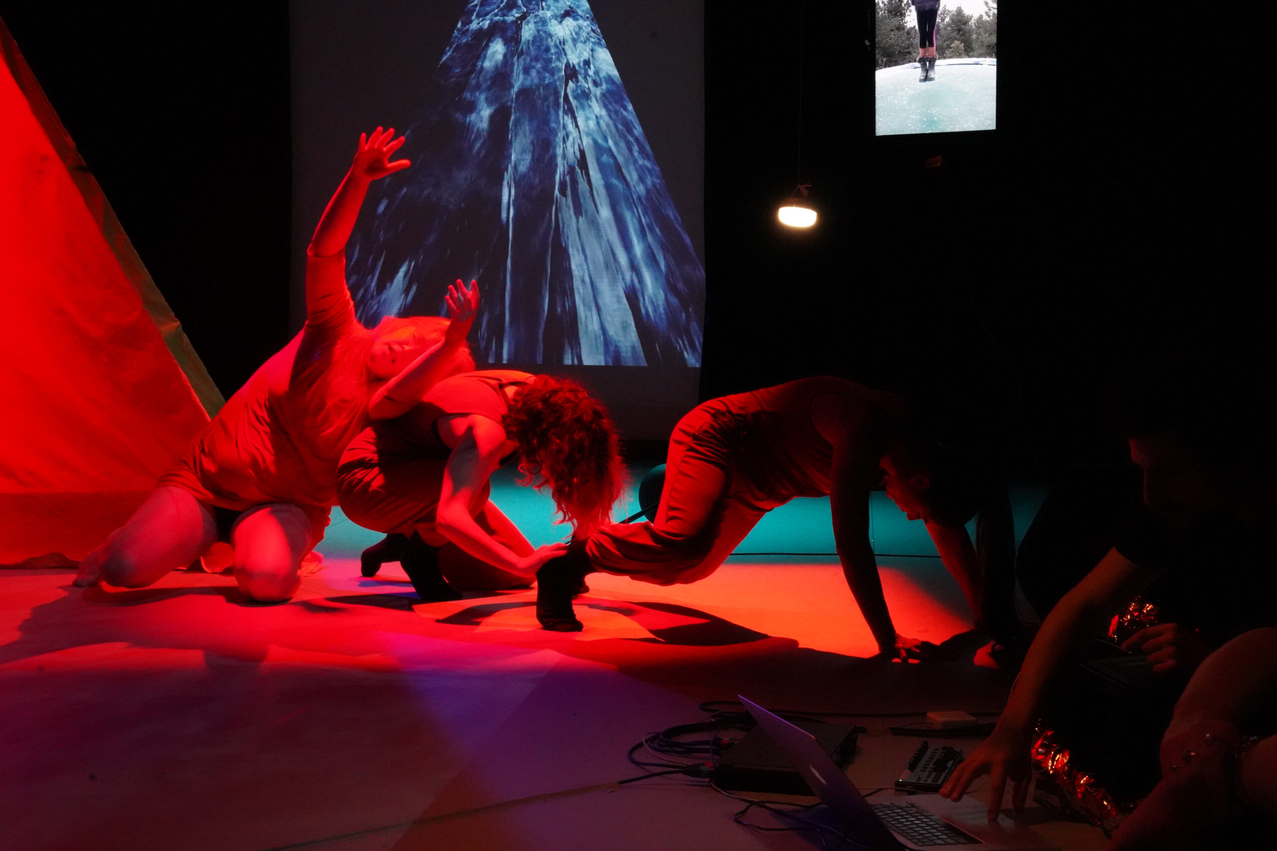 Three performers in red light are crouched in varying positions close to the ground. Behind them, blue wavey images appear on a projection mirroring the shape of an orange tent that is also in the space.