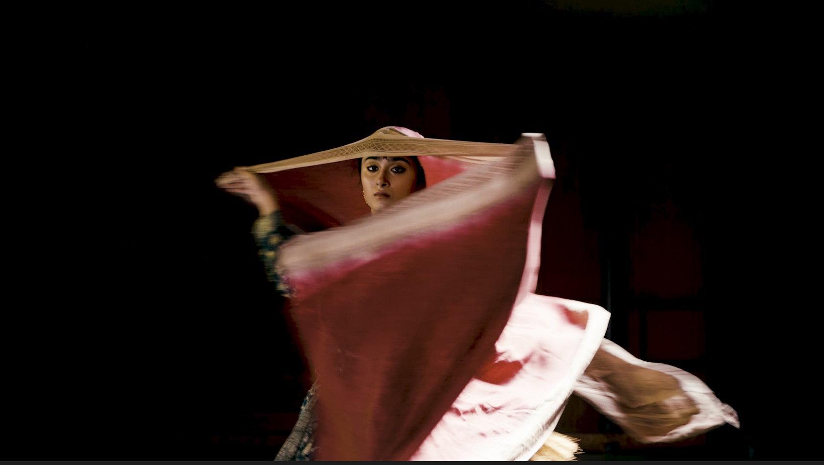 Dancer  Pradhan of Chitresh Das Dance; a dancer, against a black background, looks directly at the camera, the only part of her body visible is her face as a large piece of red and white cloth swirls around her as if she is spinning.