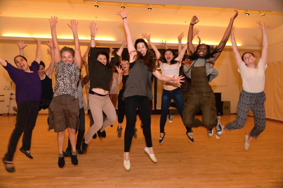 a large group of dance class students wearing different colors and styles of dance clothes, jumping in the air laughing and smiling 