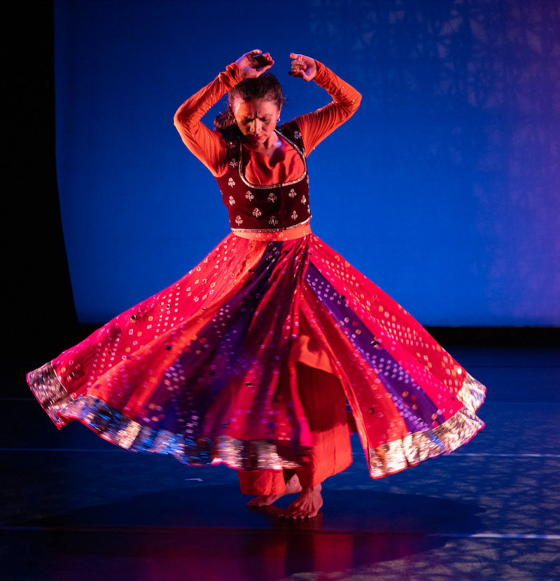 Karishma Sharma, Monsoon Dance Company stands in front of a moody blue background. They wear a red dress with a full skirt. Gaze is downward. Arms overhead.