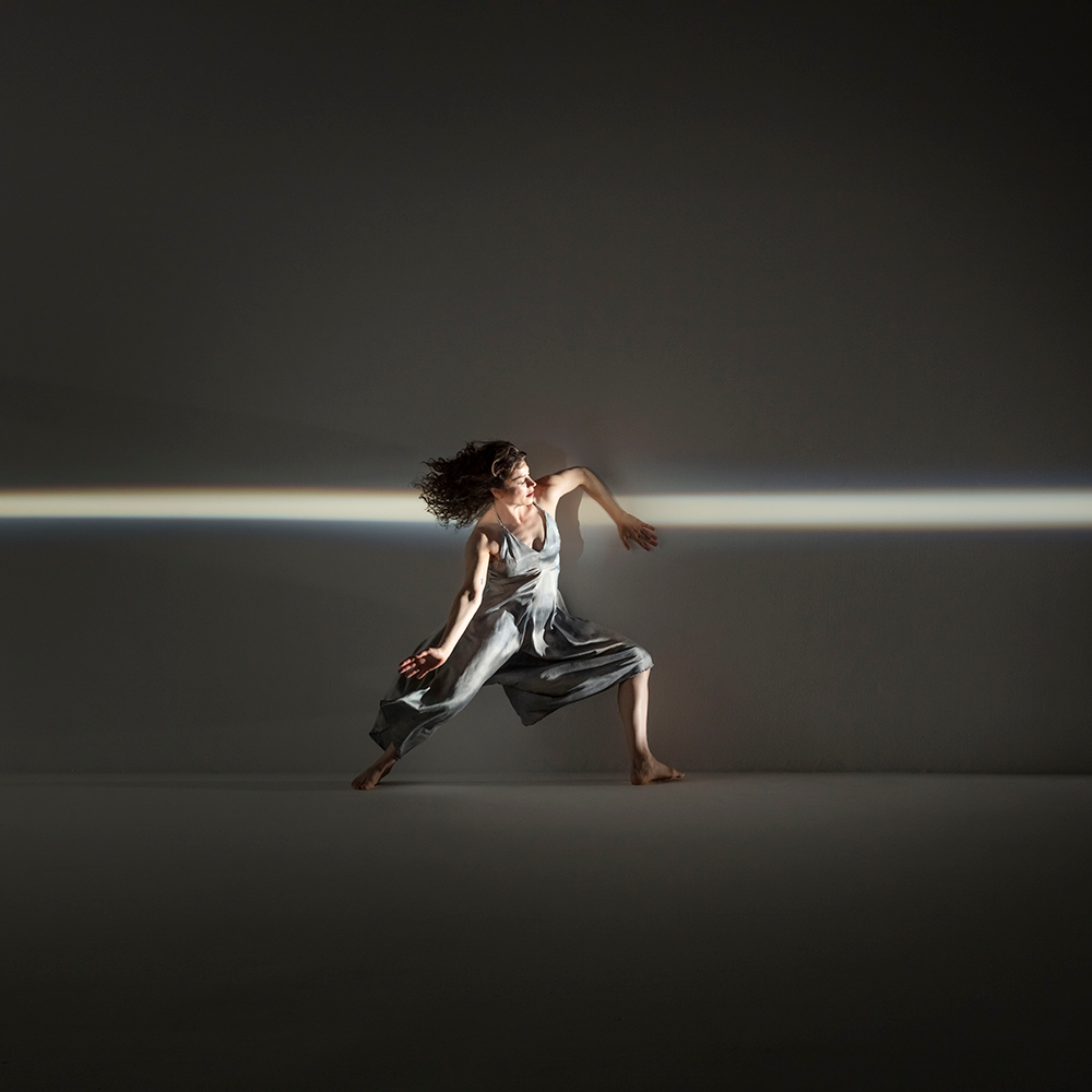 A dancer in a flowing dress performs a dynamic move under a horizontal strip of light, against a dark background. Photo by RJ Muna.