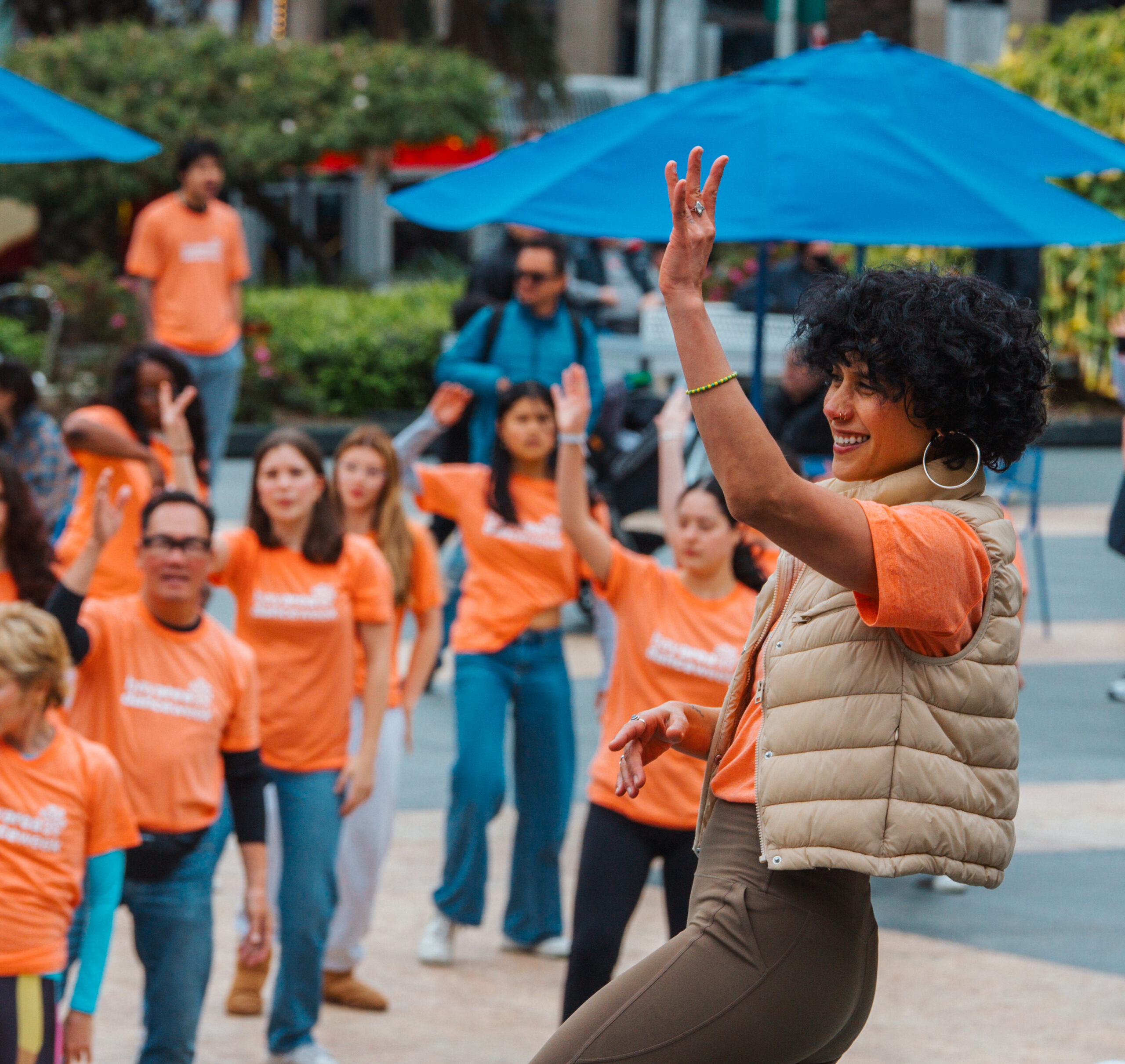 A dancer in a light brown vest with an orange shirt has their arm lifted and in the background you see others in orange shirts following along.