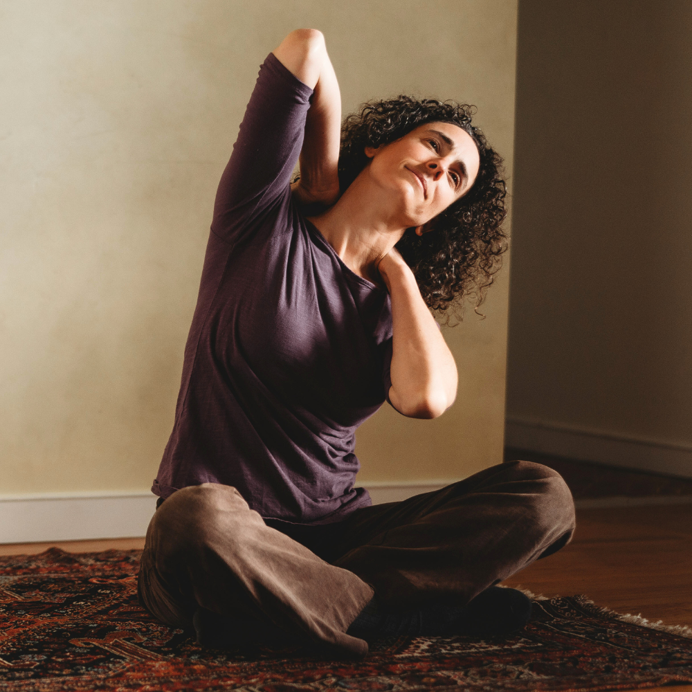 A woman sitting on a rug cross legged leaning to the left with her hands reaching from over her shoulders behind her back.