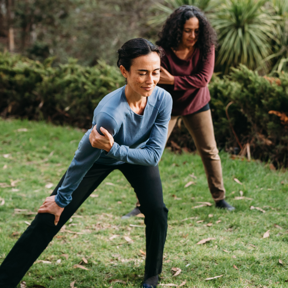 Two woman standing in the grass knees bent right leg straight body leaning to the left right hand just above the knee left hand pulling the right shoulder to the front of body.
