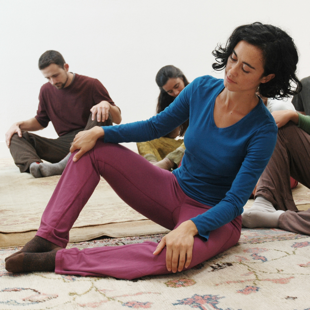 A group of people sitting on rugs with their feet sole to sole in front of them rocking back and forth with their hands on their knees.