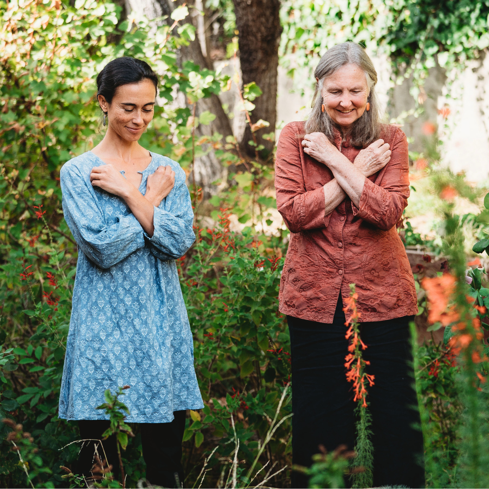 Two women standing in nature with there arms crossed over their chest one fist at each shoulder.