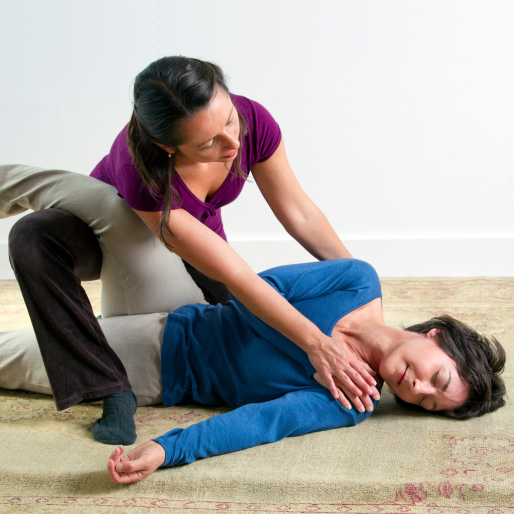 A woman kneeling on a rug performing bodywork on a woman laying with a leg draped over her knee and slightly twisted on her side..
