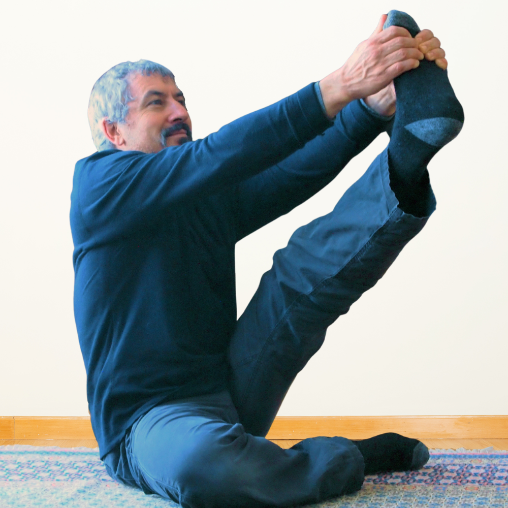 A man sitting on a rug stretching and holding his foot up in the air with his hands in front of a white wall.