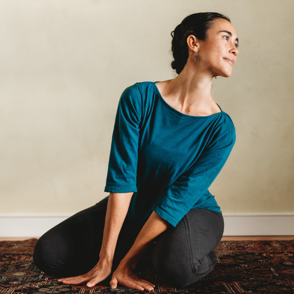 A woman kneeling on a rug body twisting and looking back over her left shoulder both hand on the floor in front supporting the twist.