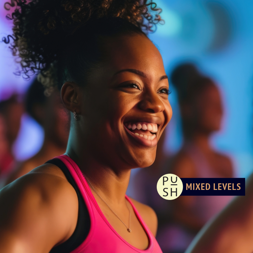 The image is a close shot of a smiling dancer with curly hair pulled back in a ponytail and wearing a hot pink tank top. The background is blurred, but bold blue and purple colors are visible as well as shadowy outlines of other dancers taking class together.