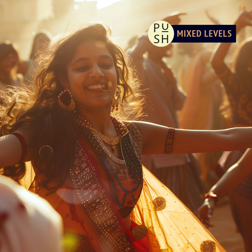 A dancer performs a Indian classical dance in a festival, smiling as others are visible in the crowd behind her. There is soft golden light shining from behind the dancers.