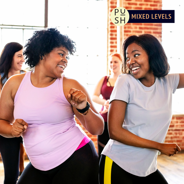 Two smiling dancers dressed in athletic wear look at each other smiling and moving their arms in the foreground, and two other dancers are visibly smiling in the background of a sunny dance studio with tall windows that extend past the frame of the image, and brick walls.