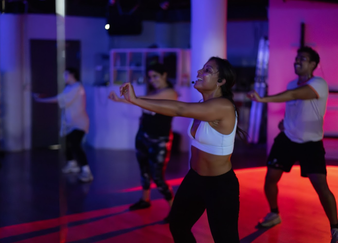 Dancers sweating and dancing together, smiling in a club-y dance class setting with dim colorful lights, all wearing glitter