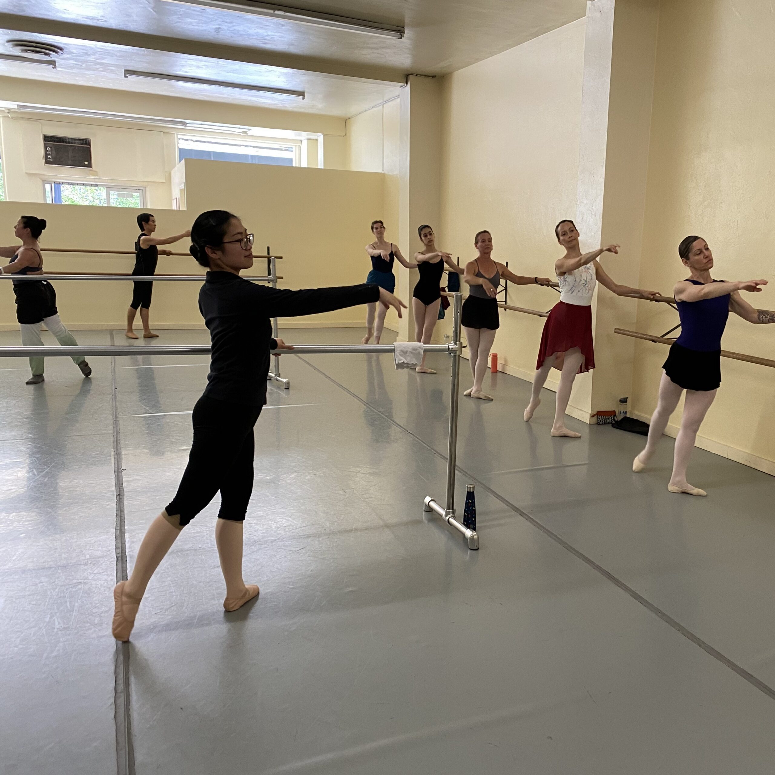 adult and teen dancers in tendu arabesque at barre taking ballet class 