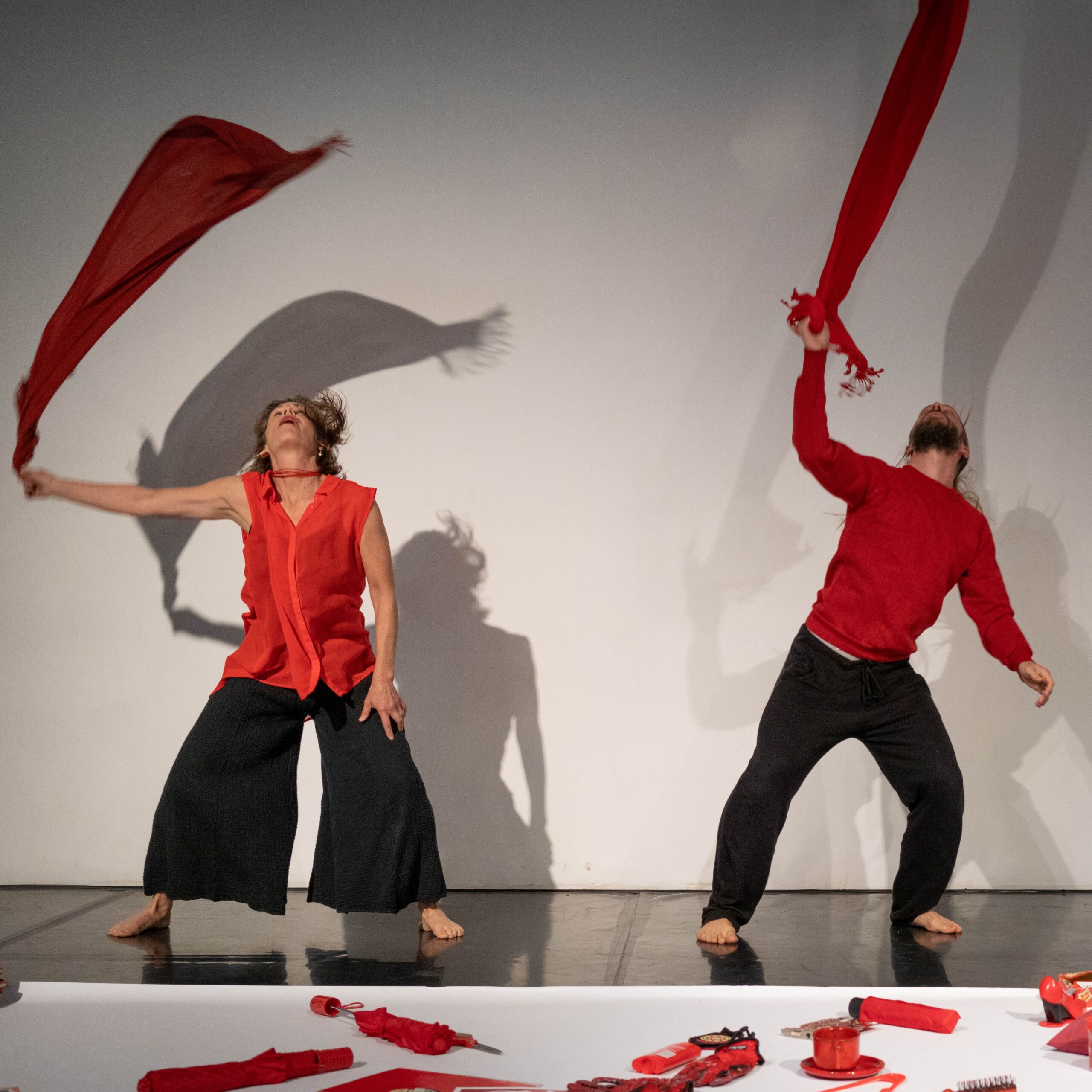 Two people dressed in red tops with black pants swirl long red scarves over their heads, so that the scarves seem suspended in the air. In front of them on the floor is a long white paper with red objects on top.
