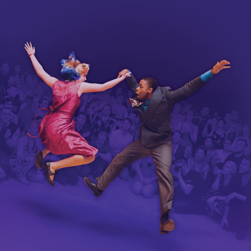 A crowd watches and cheers as a woman in a pink dress and a man in a suit leap mid-air during an energetic partner dance performance on a wooden floor.