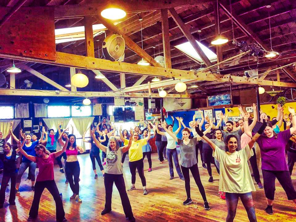 Group Bhangra dance class in a bright studio, participants dancing with raised arms and smiling.