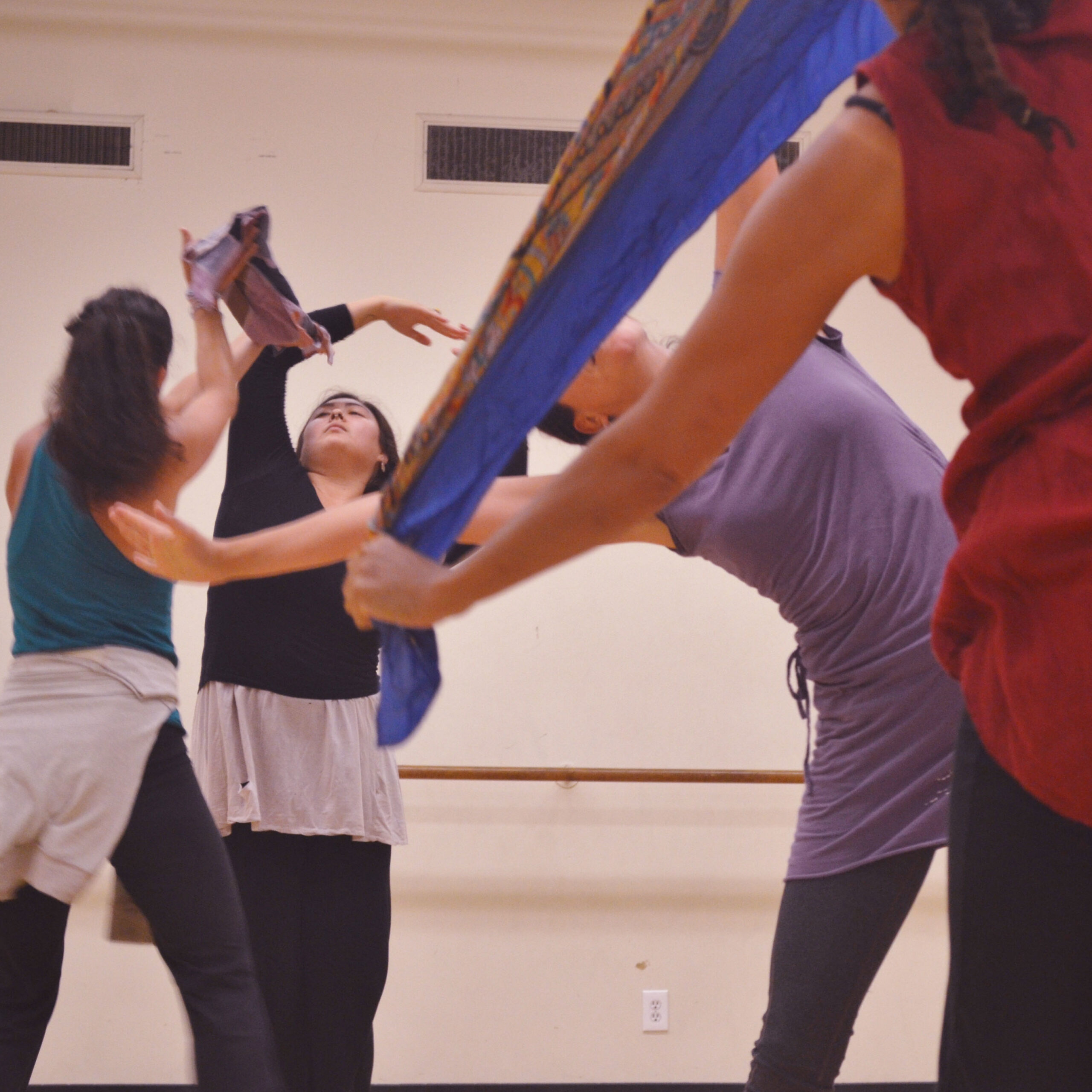 Four dancers with scarves. Two dancers in foreground face away from viewer in side bend with scarves stretched overhead, one dancer in background faces away from viewer with scarve held overhead, one dancer faces viewer with one arm curved overhead.