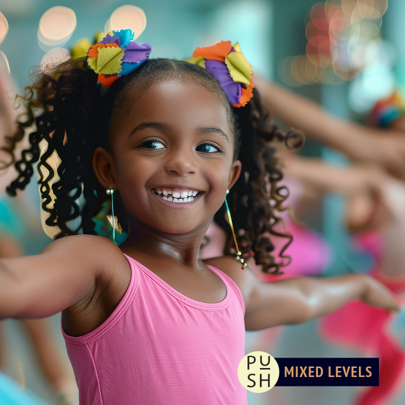 A child wearing a leotard and colorful hair ties in two ponytails smiles with their arms outstretched to the side. Other dance students are partially visible in the blurred colorful background.