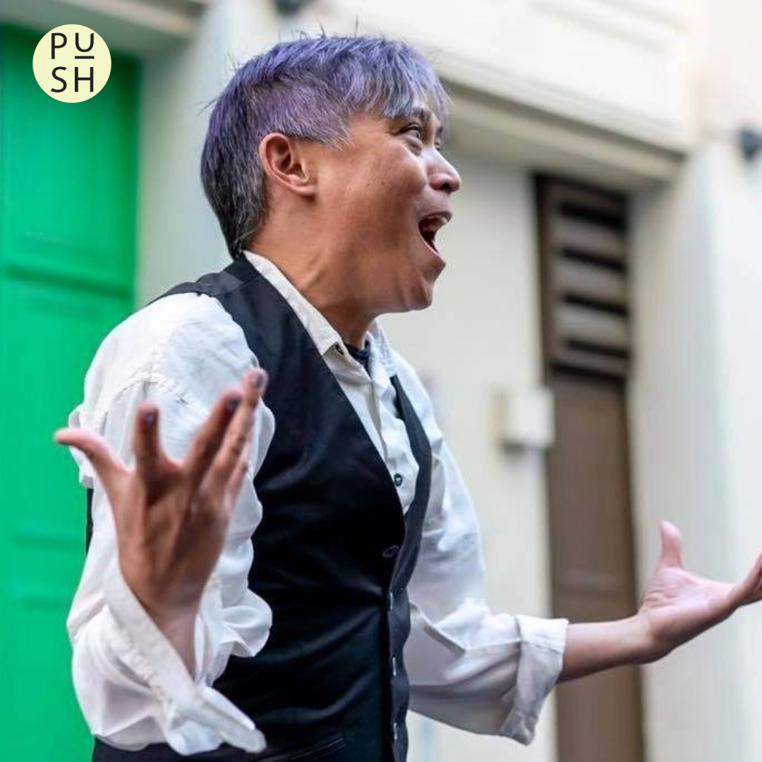 Jesse stands angled to his left, gesturing with arms out to his sides, palms facing up and fingers outstretched. Wearing a white collared shirt and black vest against a white building with a green door visible on the left, he is energetically speaking to someone not visible in the shot.