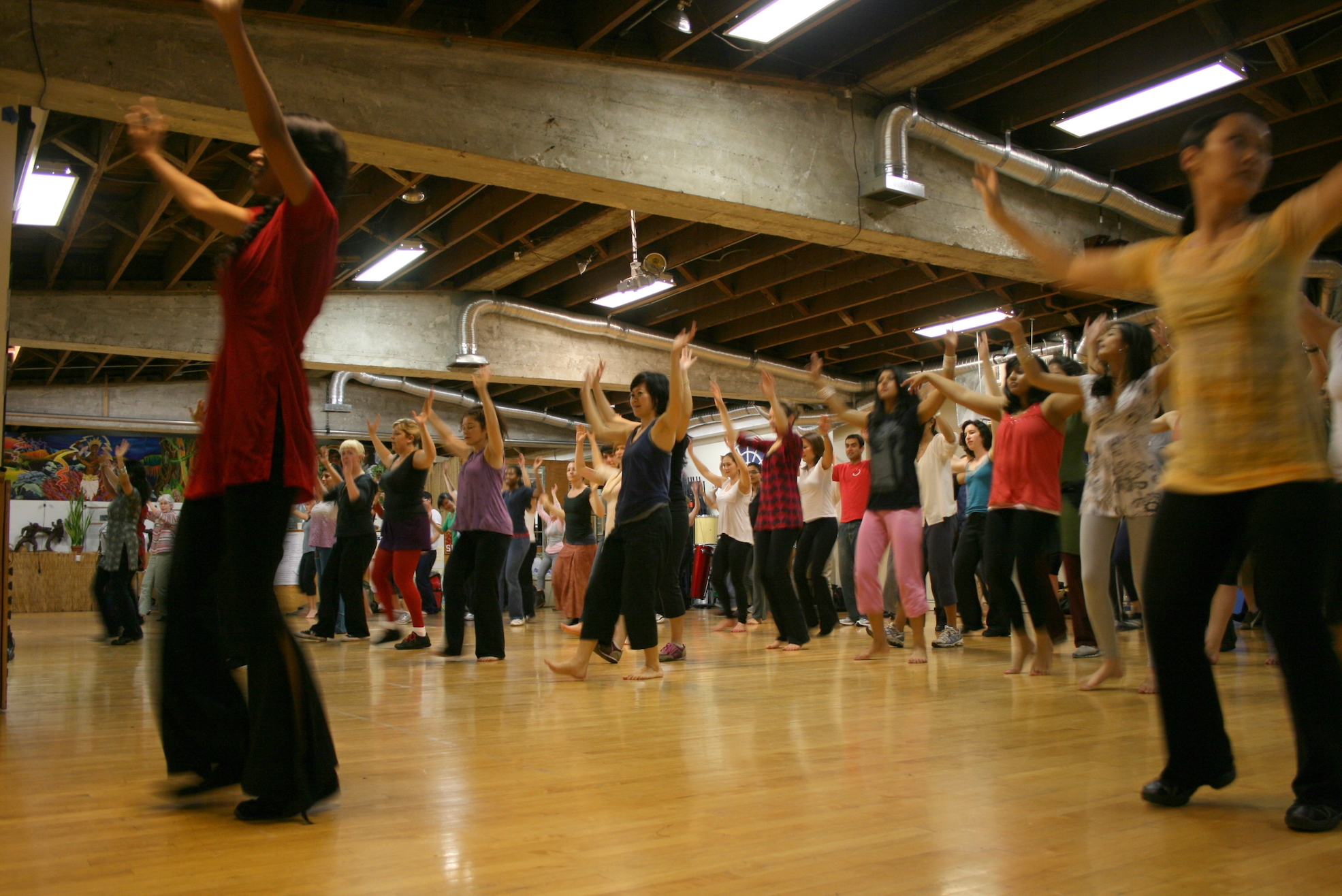 Group Bhangra dance class in a bright studio, participants dancing with raised arms.