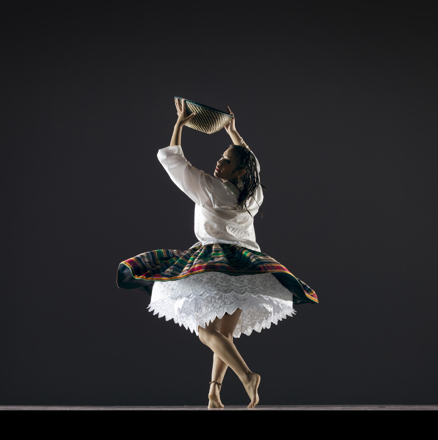 Barefoot dancer dressed in Saya Afro Boliviana costume. She is carrying a basket over her head. She is wearing a white blouse and black skirt with colorful designs.