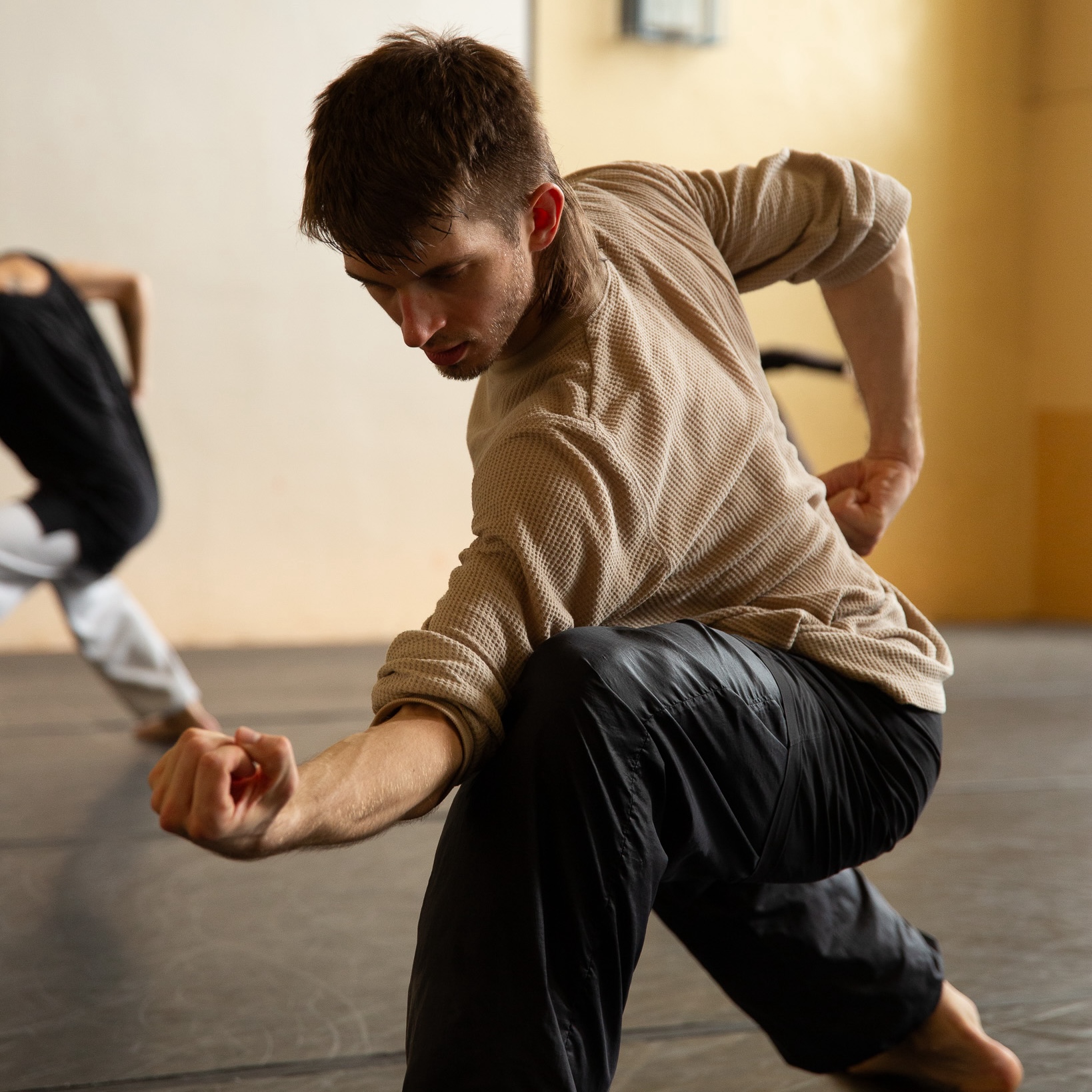 Nat Wilson wears a beige long-sleeve shirt and black pants. They perform a dynamic dance move, lunging forward with a focused expression and outstretched arm in a dance studio.