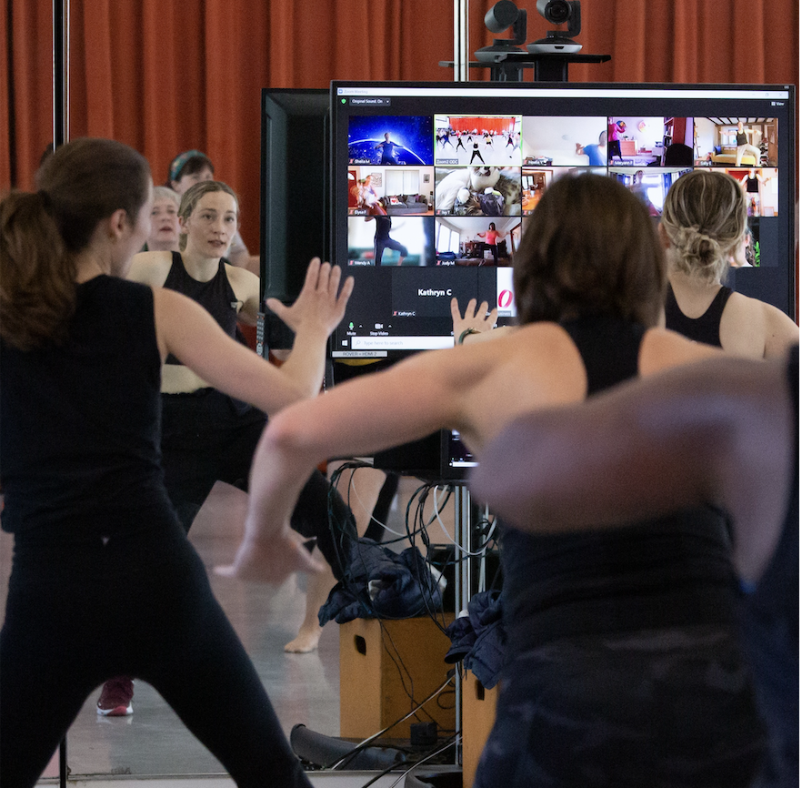 Dancers in a studio dance while facing a monitor showing a Zoom class grid of remote participants dancing at home. A hybrid R&M in-person and online dance class in progress.