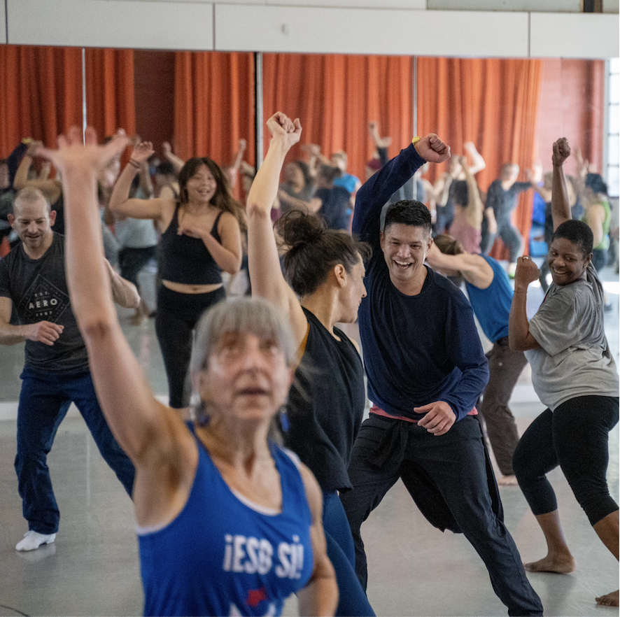 A diverse group of people of varying ages dance together joyfully in a mirrored studio, arms raised and smiling, during a Rhythm & Motion class at ODC Dance Commons in San Francisco.