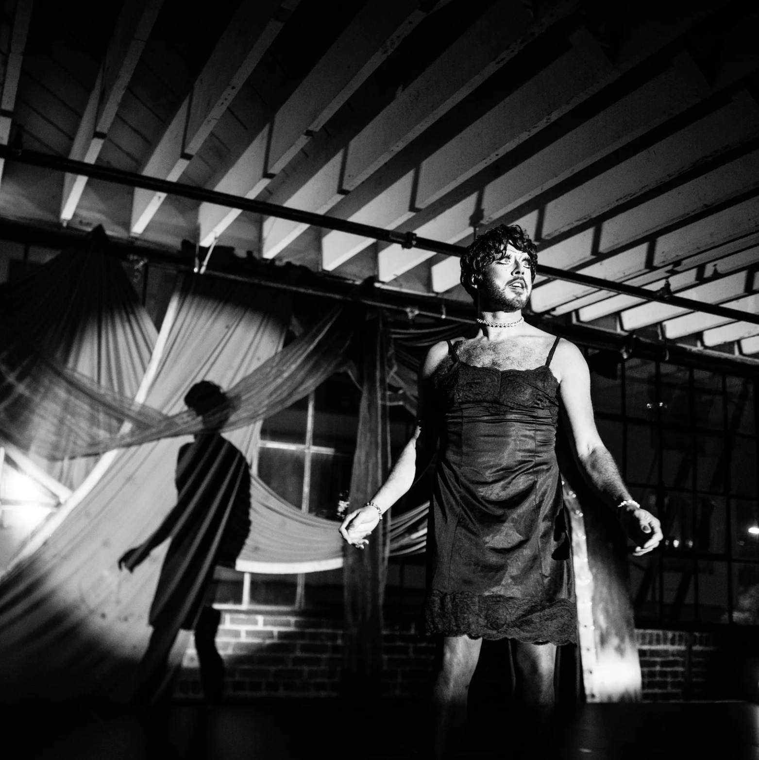 A black-and-white photo of a performer in a slip dress standing under dramatic lighting, mid-gesture, with their shadow cast against draped fabric and a windowed industrial backdrop.