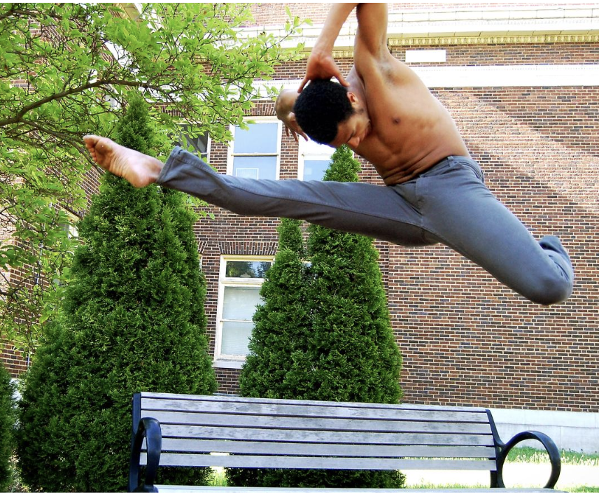 dancer jumps with one leg straight and one bent. Head is tucked and arms are bent behind their head. They look down