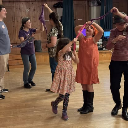 Two children ages 8 and 11 dance together with a ribbon, two adults behind them dance with a ribbon. Everyone is smiling while they dance. 
