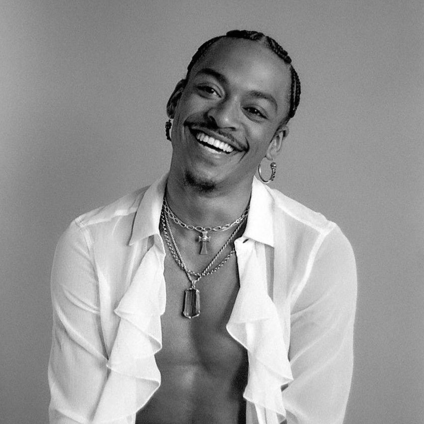 Black and white headshot of Ja'Moon who smiles while wearing hoop earrings, layered necklaces, and an open sheer shirt, posing against a plain background.