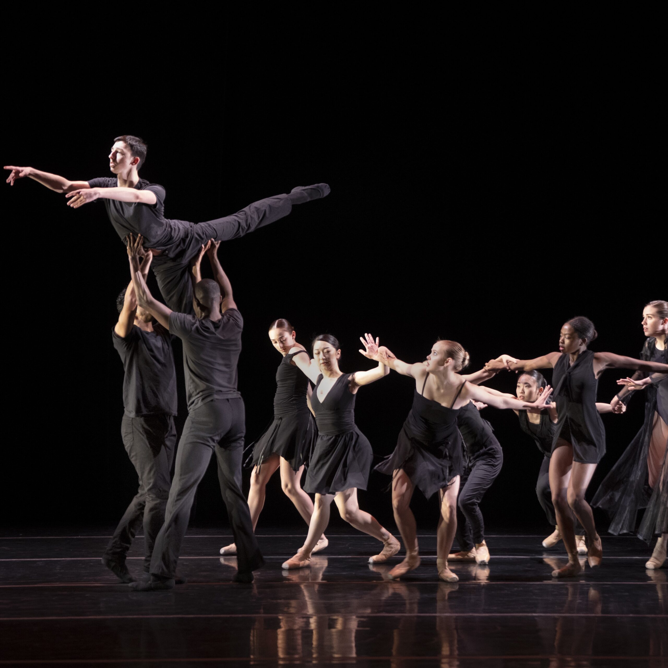 LINES Ballet Summer Program dancers perform onstage in black costumes, one lifted high into the air, reaching forward in an arabesque, by two others, as a group follows and watches.