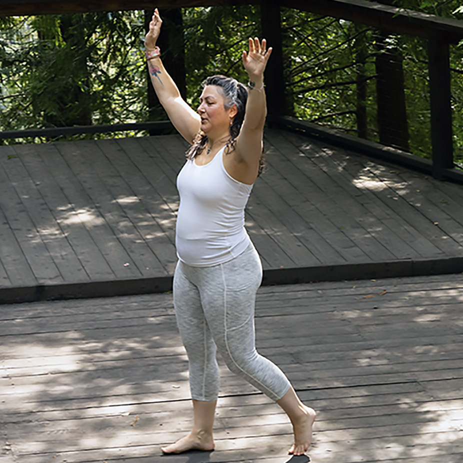 Woman with long gray hair and white shirt and gray pants reaching up to the sky with eyes closed on a wooden deck in the redwood forest