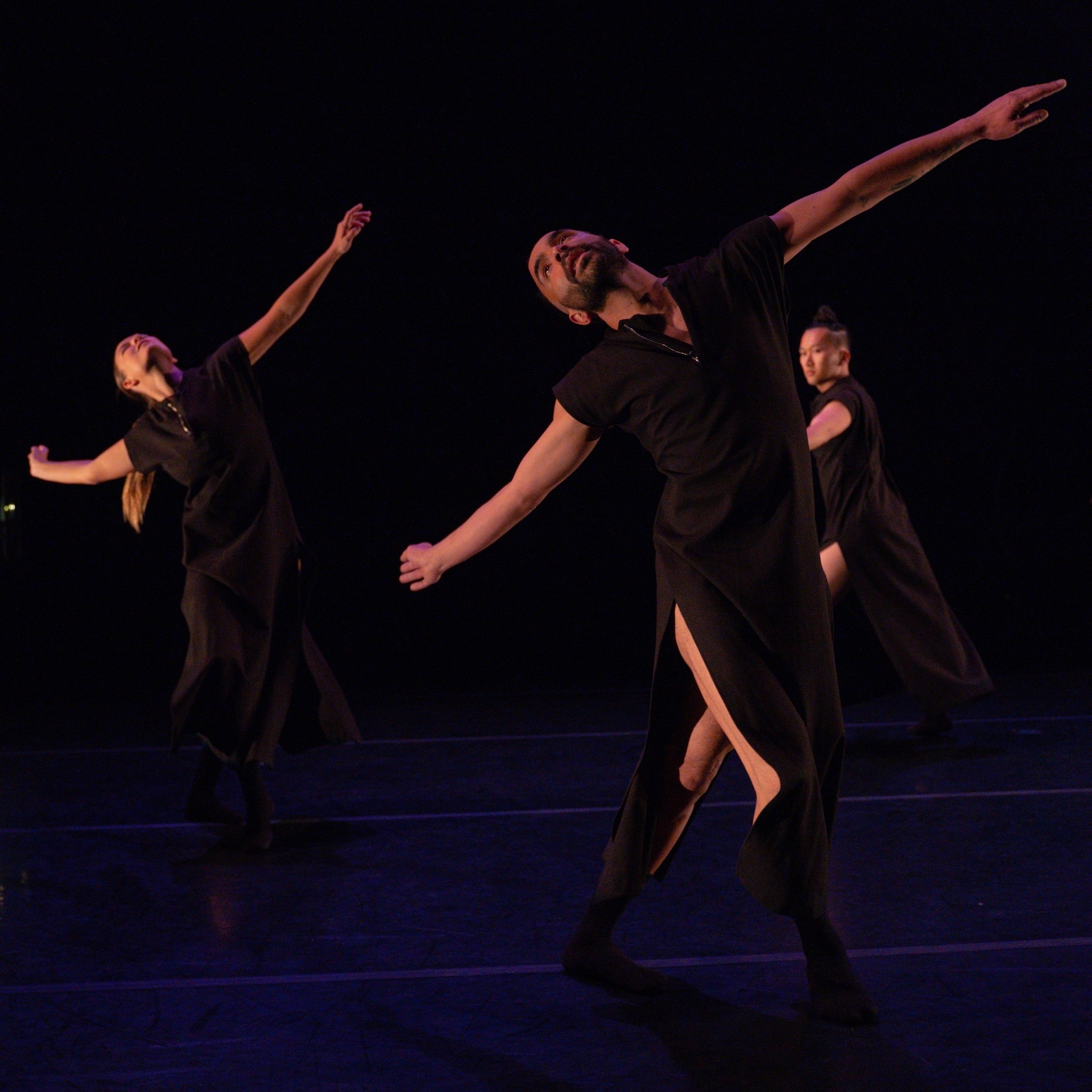 Two dancers in the foreground lean to the right with their arms outstreached to each side, their heads leaning back looking upwards.