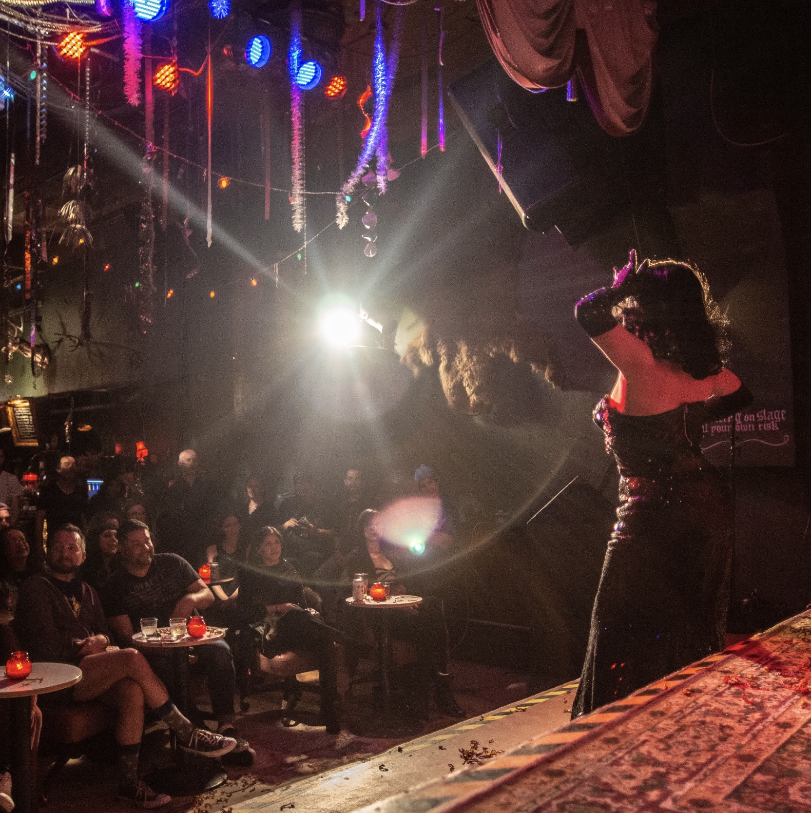 A drag performer stands bathed in stage lights, silhouetted against a glowing spotlight as they captivate an intimate audience in a cozy cabaret. Glittering décor hangs overhead, creating a warm, theatrical atmosphere filled with anticipation and applause.